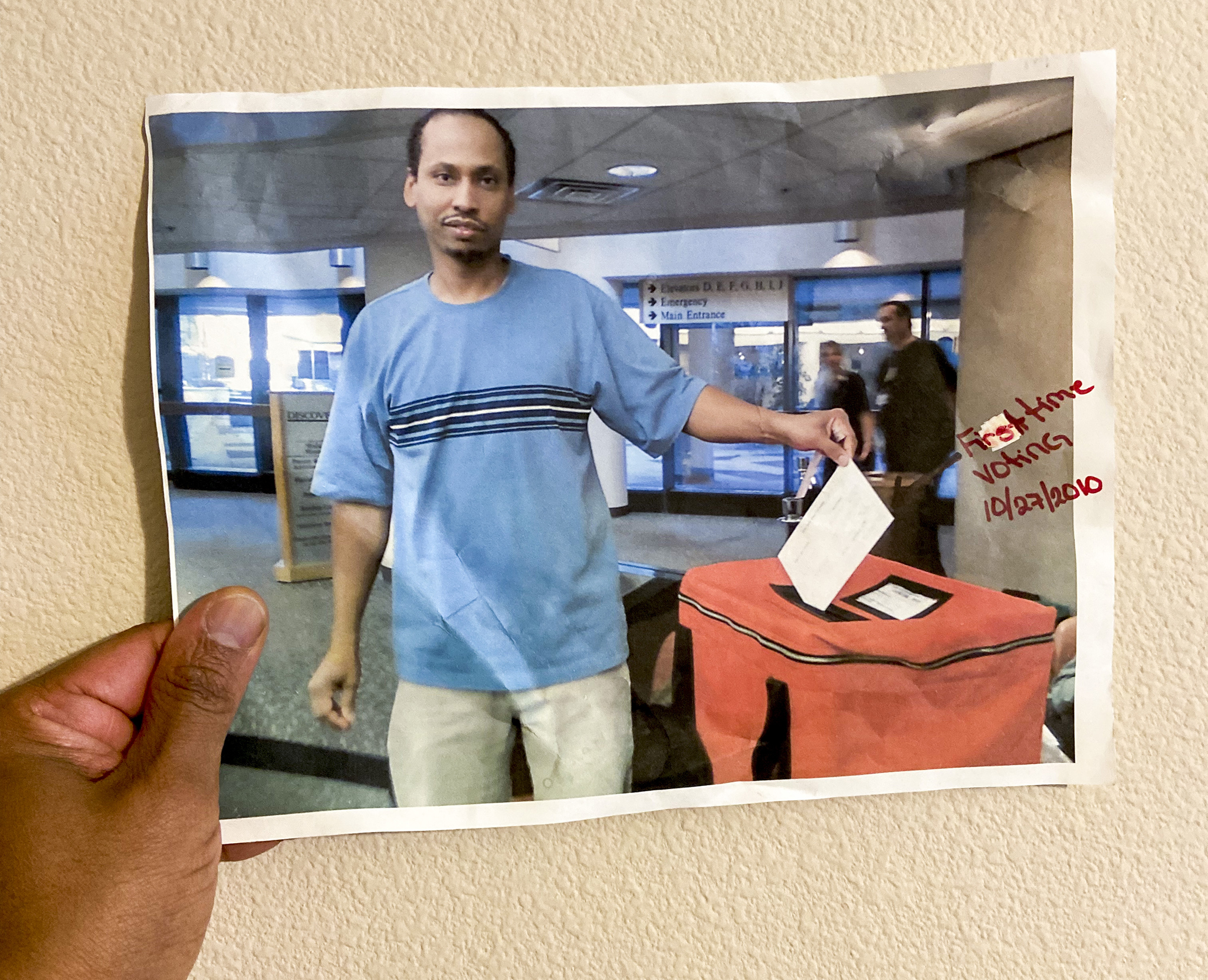 Hazim Mohamed holds an archived photo from 2010. Handwritten in red pen on top of the photo, it reads, ‘First time voting 10/27/2010.’ In the photo Hazim wears a blue striped shirt and drops a ballot into a red ballot collection box.