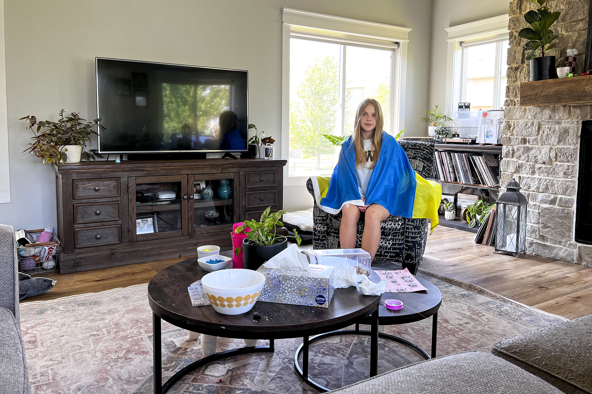 Liza sits in a chair in her host family’s living room. To her right is their TV, in front of her is a coffee table with various household objects like tissues, snacks, a house plant and friendship bracelets. Liza is wearing a gray sweatshirt with a Ukrainian flag draped around her shoulders.