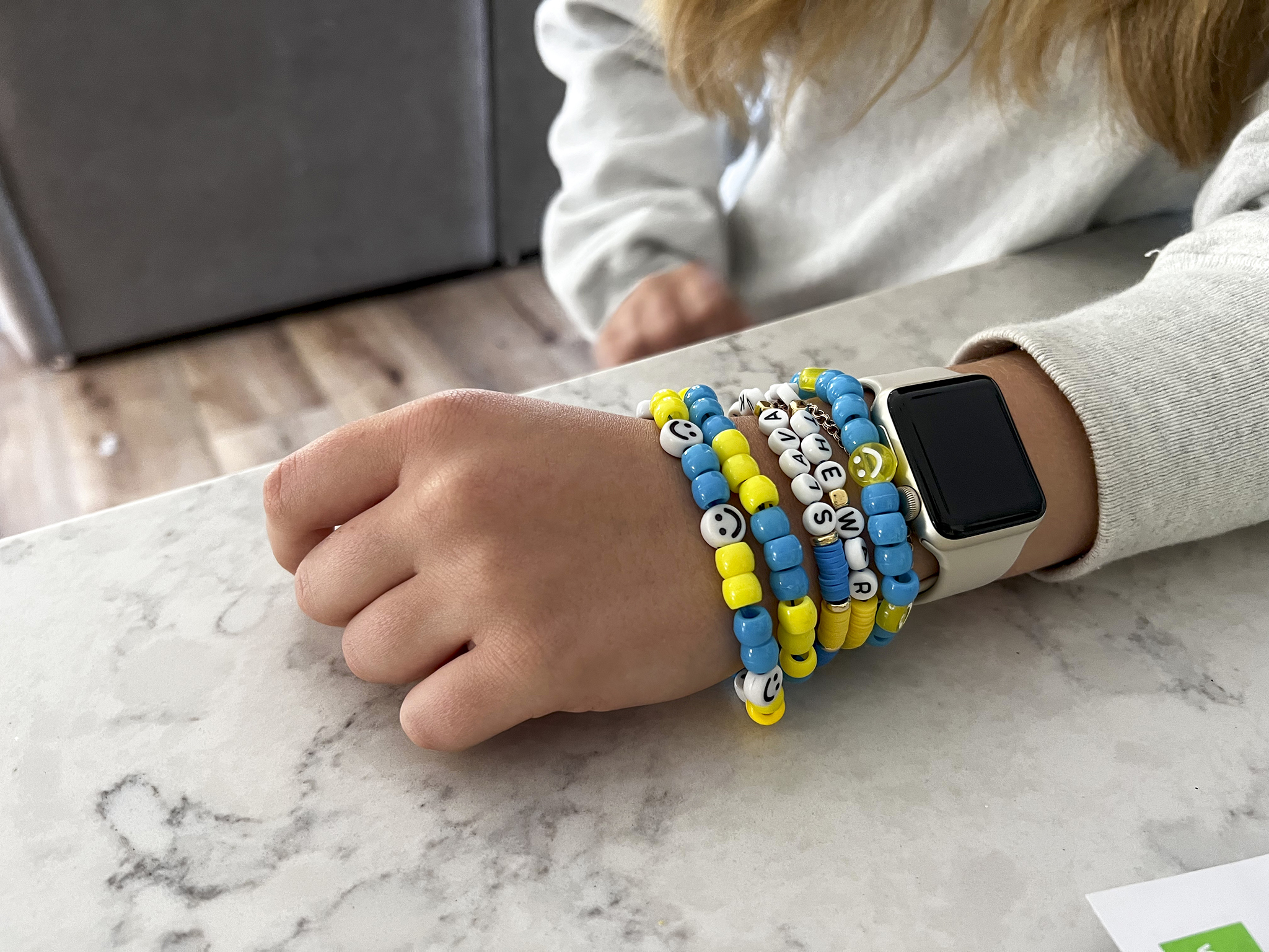 Liza’s forearm is laid flat on a granite countertop. She is wearing five bracelets with blue, yellow and white beads, an Apple Watch, and a gray sweatshirt.