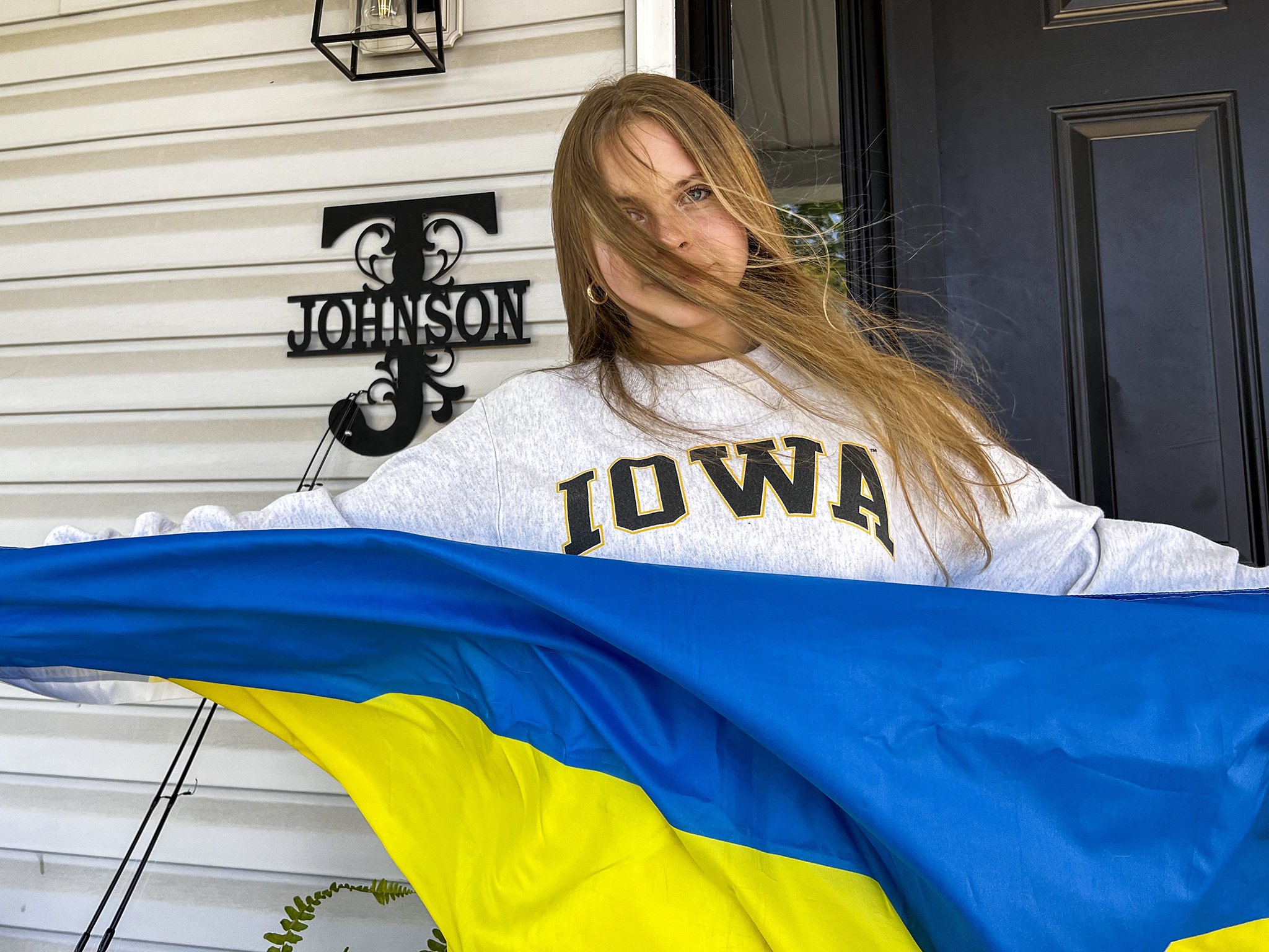 Liza stands outside her host family's front door, spanning a Ukrainian flag across her wingspan. Her hair in the wind partially covers her face. She is wearing a gray sweatshirt that reads “Iowa” across her chest. A metal “J” that reads “Johnson” hangs on the wall.