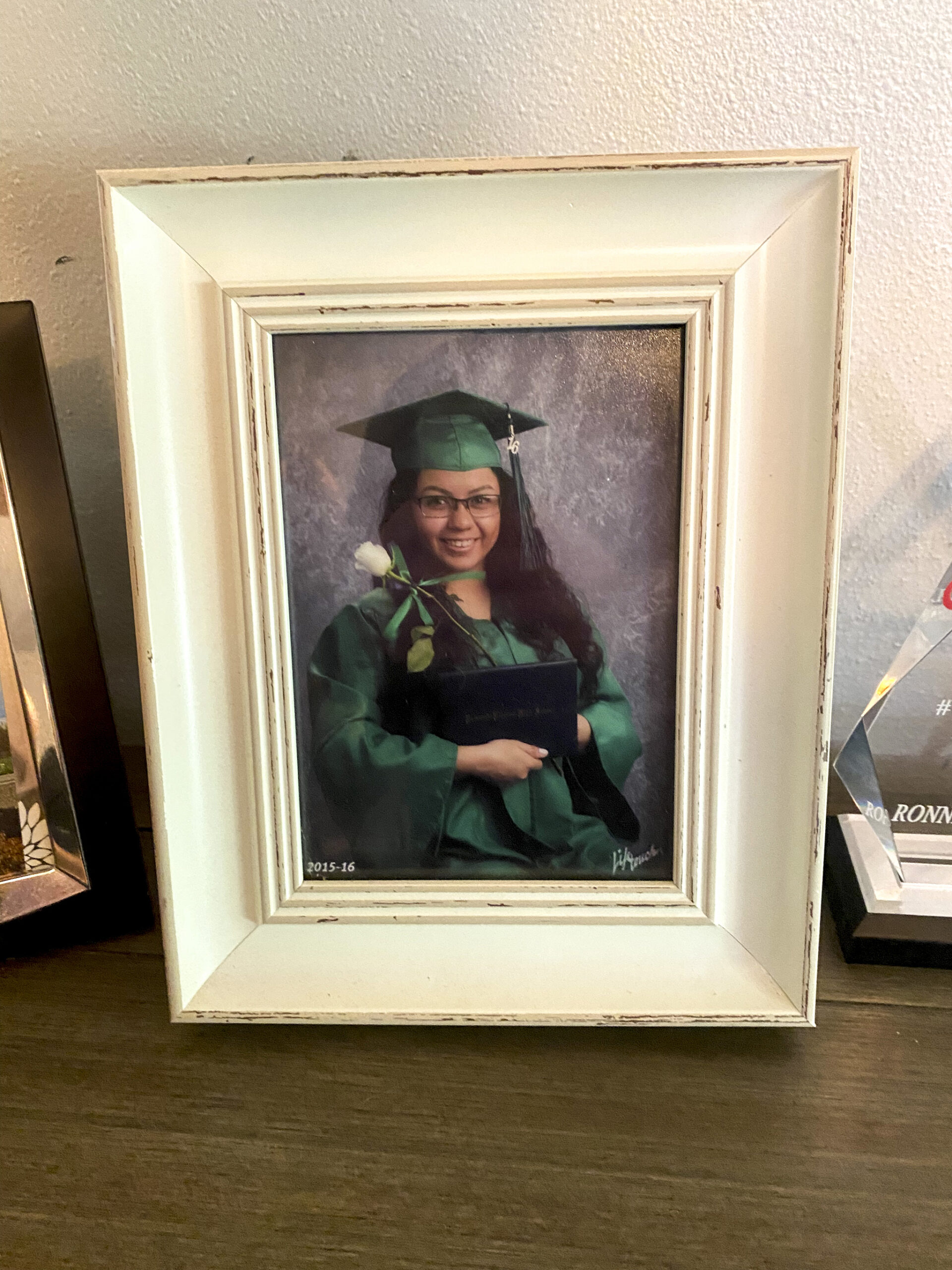 A white wooden frame contains a picture of teenage Iran Carlos-Martinez in green graduation robes. She holds a white robe and a blue leather diploma.