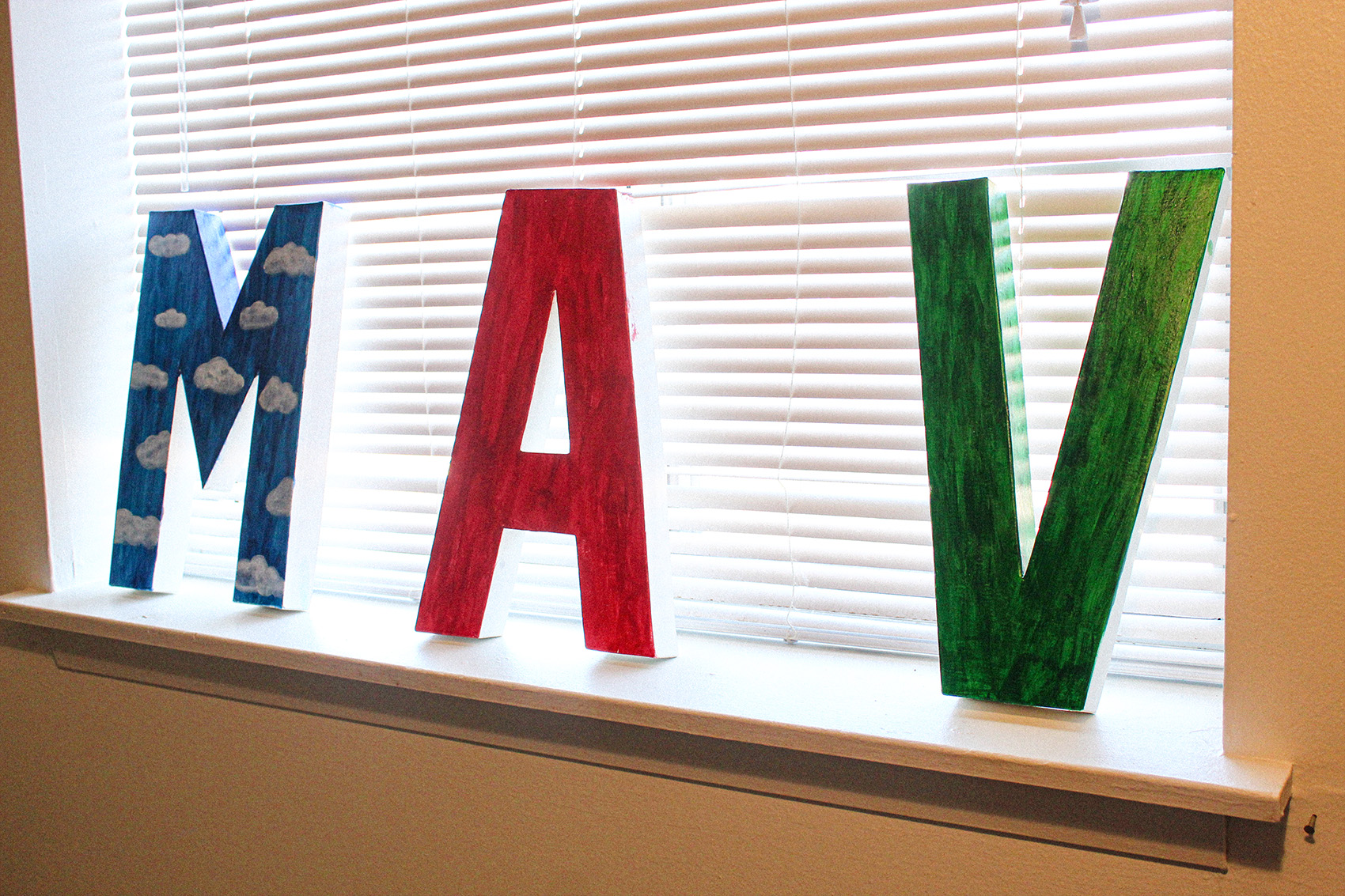 Letters (left to right) M (Blue) A (Red) and V (Green) lean against closed blinds in window.
