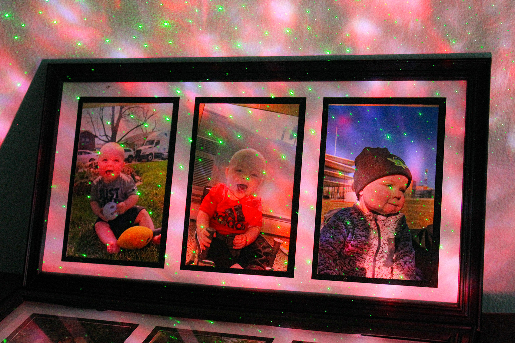 Three pictures of a baby in a 3-photo frame leaning against a wall with pink and purple light with green dots shining over it.
